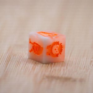 Puede incluir: Un dado cuadrado blanquecino con ilustraciones naranjas de un león, un mono y otros animales. El dado está sobre una superficie de madera de color marrón claro.