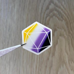 May include: A geometric sticker in the shape of a 12-sided die, featuring the non-binary pride flag colours: yellow, white, purple, and black. The sticker is held by tweezers against a wooden background.