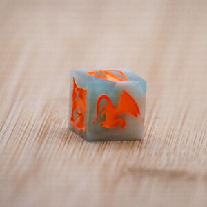 Könnte beinhalten: Ein quadratischer, handgefertigter Würfel mit einem Drachendesign. Der Würfel ist aus hellblauem und klarem Harz mit orangefarbenen Drachensilhouetten. Der Würfel liegt auf einer Holzoberfläche.