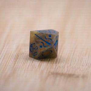 May include: A six-sided, polyhedral die with a golden-brown hue and blue markings. The die is made of a translucent material, with the blue numbers and symbols visible inside. The die is designed for tabletop gaming.