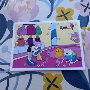 May include: A rectangular art print featuring a colorful cartoon scene with characters Marceline, Princess Bubblegum, Jake, and Finn playing a card game on a pink floor. The background includes potted plants, a shelf, and a pizza-eating character.