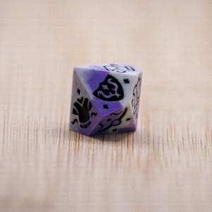 Pode incluir: Um dado de várias faces, roxo e branco marmoreado, com símbolos pretos. O dado está sobre uma superfície de madeira castanha clara. Os símbolos incluem um escudo, uma mão e outros desenhos abstratos.