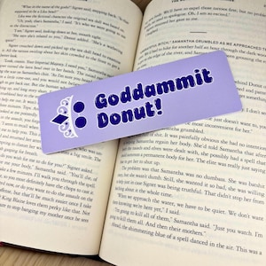 May include: A rectangular light purple bookmark featuring the dark blue text Goddammit Donut! in a bold, rounded font. To the left of the text is a white graphic of a tiara with a small blue diamond shape centred at the base of the crown.