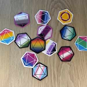 Op de afbeelding: Een verzameling kleurrijke, geometrische stickers met verschillende pride flag ontwerpen. Elke sticker heeft een veelzijdige vorm met een witte omtrek en een zwarte rand. De stickers zijn gerangschikt op een lichtbruin houten oppervlak.