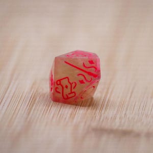 Pode incluir: Um dado translúcido de várias faces com marcações rosa. O dado tem um interior bege claro e símbolos e números rosa em cada face. O dado está sobre uma superfície clara com grão de madeira castanha.