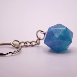 Puede incluir: Un llavero con un dije geométrico azul y turquesa. El dije está unido a una cadena y un llavero plateados. Este llavero es un accesorio decorativo.