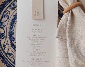 May include: A wedding menu card with a beige tag tied with a white ribbon. The menu is on a white card, with a list of dishes. The card is on a blue and white patterned plate, with a beige napkin and wooden ring.