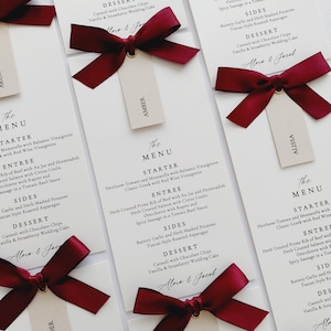 Może przedstawiać: Eleganckie karty menu weselnego z bordowymi kokardami i etykietami z imionami. Karty zawierają menu z opcjami przystawek, dań głównych, dodatków i deserów. Menu zawiera dania takie jak pieczeń wołowa w ziołach i cannoli.