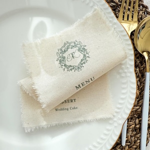 Op de afbeelding: Twee beige linnen menukaarten met gerafelde randen, bedrukt met een groene bloemenkrans en de letter "K". Het woord "MENU" staat onder de krans gedrukt. De onderste kaart leest "DESSERT" en "Wedding Cake".