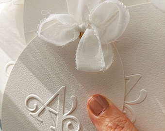 May include: White round paper card with a raised, embossed letter "A" in the center. The card is decorated with a white sheer ribbon tied in a bow.