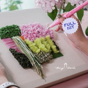 Op de afbeelding: Een close-up van iemand die een roze haaknaald gebruikt om een ​​kleurrijk garenwandkleed te maken. De wandkleed heeft verschillende texturen en kleuren garen, waaronder groen, roze, geel en wit. Het canvas is gelabeld met "tiny hand" en een sticker met de tekst "FULL KIT".