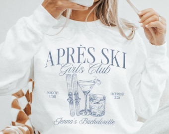 May include: White long-sleeve sweatshirt with a blue graphic that reads "APRÈS SKI Girls Club" with ski equipment and a cocktail illustration. The text "Jenna's Bachelorette" is also included. The sweatshirt is paired with gray sweatpants.