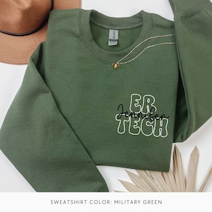 May include: A military green sweatshirt with the text "ER Jennifer TECH" in white outlined letters. The sweatshirt is folded and laid on a white surface with a brown hat and a dried palm leaf.