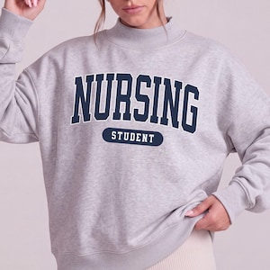 May include: A light gray crewneck sweatshirt with a mock neck collar and long sleeves. The front features the dark navy blue text NURSING in a collegiate font, with the word STUDENT printed in white inside a dark navy blue horizontal oval.