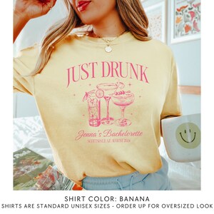 May include: A yellow t-shirt with the text "JUST DRUNK" in pink lettering. The shirt also features a pink illustration of three cocktails. The text "Jenna's Bachelorette Scottsdale AZ August 2024" is printed below the illustration. The shirt colour is described as "BANANA".