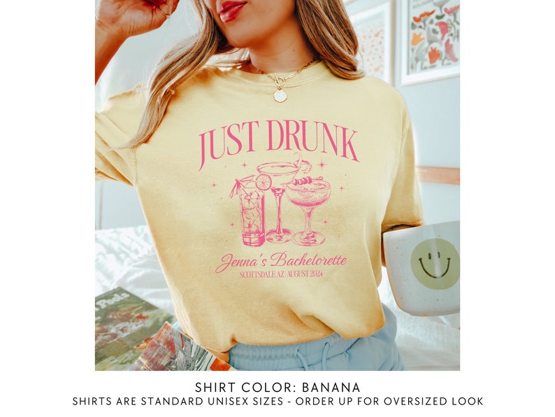May include: A yellow t-shirt with the text "JUST DRUNK" in pink lettering. The shirt also features a pink illustration of three cocktails. The text "Jenna's Bachelorette Scottsdale AZ August 2024" is printed below the illustration. The shirt colour is described as "BANANA".