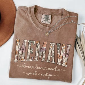 May include: A brown t-shirt with the word "MEMAW" in large letters with floral designs. The names "olivia", "liam", "amelia", "jacob", and "evelyn" are written below the word "MEMAW" with hearts between each name.