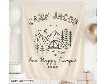 Puede incluir: Bandera de tela color marfil con el texto verde "CAMP JACOB" y "One Happy Camper". La bandera presenta una escena de campamento con montañas, árboles y una tienda de campaña. La bandera está decorada con globos y regalos.