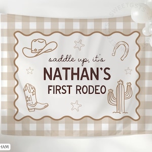 May include: Tan gingham backdrop with a scalloped border featuring cowboy-themed illustrations. The text reads "saddle up, it's NATHAN'S FIRST RODEO" in brown lettering. Includes a cowboy hat, horseshoe, cactus, and cowboy boot.