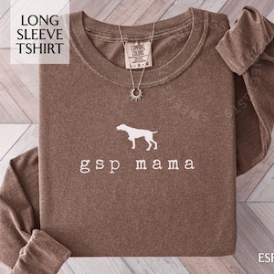 May include: Espresso-colored long sleeve t-shirt with the text "gsp mama" and a white silhouette of a dog. The shirt has a crew neck and a relaxed fit. A silver necklace with a sunburst pendant is visible.