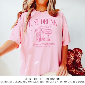 May include: Pink t-shirt with the text "JUST DRUNK" and a graphic of three cocktails. The text "Jenna's Bachelorette Scottsdale AZ August 2024" is below the graphic. The shirt colour is blossom.