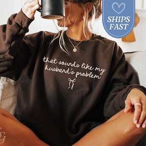 May include: A dark brown sweatshirt with the phrase "that sounds like my husband's problem" in white script. The sweatshirt has long sleeves and a crew neck. The image also includes a black mug.