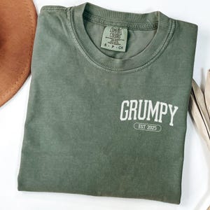 May include: Olive green Comfort Colors t-shirt with the word "GRUMPY" printed in white on the back, along with "EST 2025". The shirt has a crew neck and a small tag at the neckline.