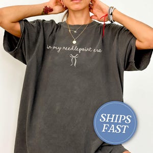 May include: Dark gray oversized t-shirt with the text "in my needlepoint era" in a script font, and a bow graphic. The shirt has a vintage, worn look. The shirt is a casual, comfortable style.
