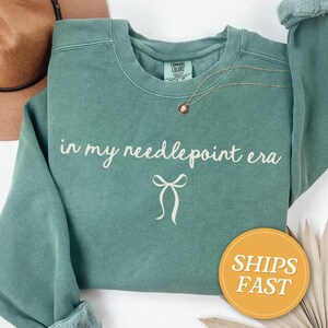 May include: A teal crewneck sweatshirt with the phrase "in my needlepoint era" in white script and a bow graphic. A gold necklace with a pendant is draped on the shirt. A brown hat is visible in the upper left corner.