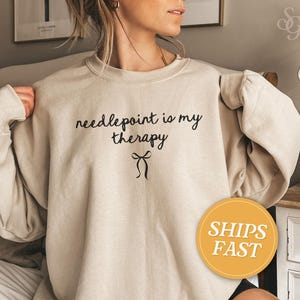 May include: A beige sweatshirt with the phrase "needlepoint is my therapy" in black script, along with a bow graphic. The sweatshirt has long sleeves and a crew neck.