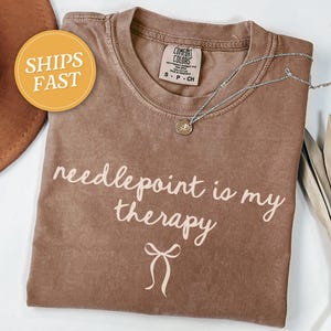 May include: A brown t-shirt with the phrase "needlepoint is my therapy" in white script. A silver necklace with a pendant rests on the shirt. A round sticker with "SHIPS FAST" is visible.
