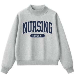 May include: A heather gray long-sleeve sweatshirt with a mock neck collar. The front features the dark blue text NURSING in a collegiate font, with the word STUDENT printed in white inside a dark blue horizontal pill-shaped graphic below it.