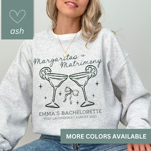 May include: A gray sweatshirt with the text "Margaritas & Matrimony" and two margarita glasses with limes. The text "Emma's Bachelorette" is below the glasses with the date and location.