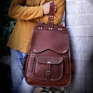 May include: A brown leather backpack with multiple pockets and adjustable straps. The backpack has a flap closure and buckle details. The bag is held by a person wearing a mustard-colored shirt and blue jeans.