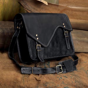 May include: A black leather messenger bag with white stitching and brass-colored buckles. The bag has a flap closure and an adjustable shoulder strap. The bag is sitting on a brown burlap surface.