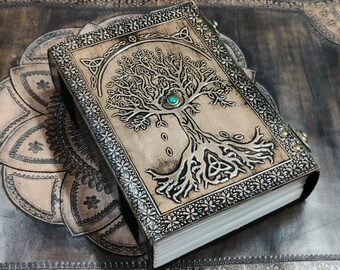 May include: A brown leather journal with a tooled design of a tree of life with a green gemstone at the center. The journal has a Celtic knot design at the base of the tree.