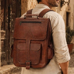 Könnte beinhalten: Ein brauner Lederrucksack mit einem Tragegriff, einer horizontalen Reißverschlusstasche auf der Klappe und einer großen Vordertasche, die mit einem vertikalen Riemen und einer Metallschnalle gesichert ist. Der Rucksack hat zwei Seitentaschen.
