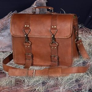 May include: A brown leather briefcase with a top handle and a detachable shoulder strap. The bag features two front straps with buckles and brass-colored hardware. The leather has a natural, textured appearance.