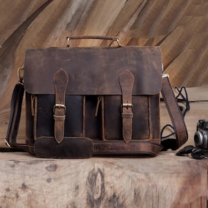 May include: A brown leather satchel with a flap closure, two buckle straps, and a top handle. The bag has a shoulder strap and a front pocket. The leather has a distressed finish, giving it a vintage look. The bag is sitting on a wooden surface.