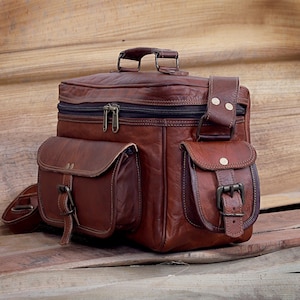 May include: A brown leather satchel with multiple pockets and a shoulder strap. The bag features a top handle, a zippered main compartment, and two front pockets with buckle closures. The leather has a distressed appearance.