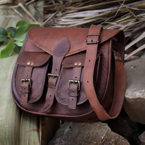 May include: A brown leather satchel bag with two front pockets and a long adjustable strap. The bag has a flap closure with buckles and metal accents.
