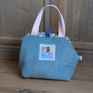 Peut inclure: Un petit sac à main en velours côtelé bleu clair avec des poignées roses. Le sac présente un écusson brodé "RAINBOW" coloré et un intérieur à motifs. Une petite languette à pois bleus ferme le dessus.