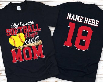 May include: Two black t-shirts. One shirt has the text "My Favorite Softball Player Calls Me Mom" with a red heart and a softball and bat graphic. The other shirt has the text "NAME HERE 18" in red.