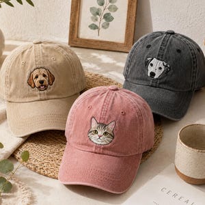 May include: Three baseball caps in tan, dusty pink, and charcoal gray. The tan cap features an embroidered golden-brown dog, the pink cap displays an embroidered tabby cat face, and the charcoal gray cap shows an embroidered white and black dog.