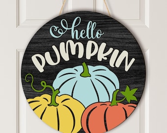 Puede incluir: Un letrero redondo de madera con un fondo de veta de madera oscura que presenta el texto "hello PUMPKIN" en letras cursivas y de molde de color azul claro y crema. Debajo del texto hay tres calabazas ilustradas en azul claro, amarillo y naranja.