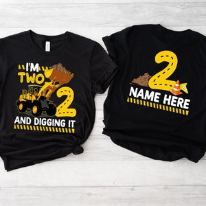 May include: Two black t-shirts with a construction theme. One shirt features a yellow excavator with the text "I'm Two and Digging It". The other shirt has a yellow number "2" with the text "Name Here".