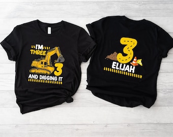 May include: Two black t-shirts with a construction theme. One shirt says "I'm Three and Digging It" with a yellow excavator and the number 3. The other shirt says "Elijah" with the number 3 and a construction cone.