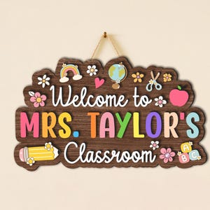 Puede incluir: Letrero de aula de madera con el texto "Welcome to MRS. TAYLOR'S Classroom". El letrero presenta letras coloridas y elementos decorativos como un arco&iacute;ris, un globo terr&aacute;queo, una manzana y flores. Est&aacute; dise&ntilde;ado con un fondo de vetas de madera y una cuerda para colgar.