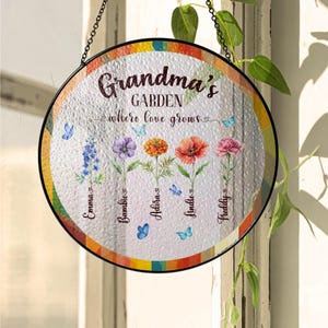 Puede incluir: Un letrero de jardín redondo de vidrio colorido con el texto "Grandma's Garden where love grows" y cinco flores con nombres escritos debajo: Emma, Bombie, Alora, Lindle y Freddy.