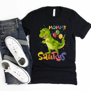 May include: Black t-shirt featuring a green dinosaur holding colourful balloons. The text "Mommy Saurus" is written in rainbow colours.